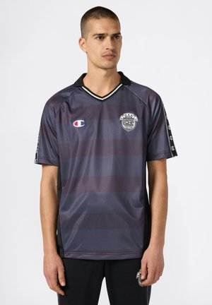 Hombre vestido con una camiseta deportiva de rayas oscuras con logos en el pecho y las mangas, de pie con una expresión neutra frente a un fondo sencillo.