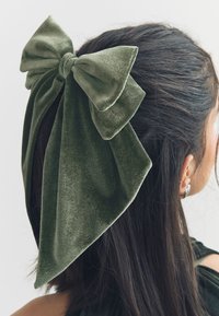 Velvet hair bow in dark green, featuring a large, prominent design with textured fabric and flowing tails, secured at the base with a knot.
