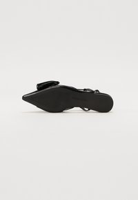 Black patent leather high-heeled shoe with a pointed toe, featuring a strap and a textured sole for grip.