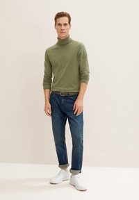 Pull à col roulé vert, jeans en denim bleu et baskets blanches. Le pull a une coupe ajustée ; les jeans sont retroussés au niveau des chevilles, look décontracté.