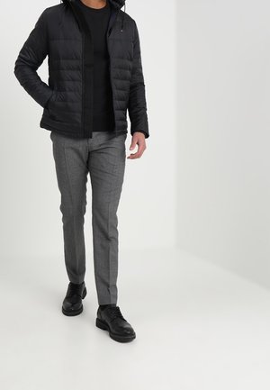 Chaqueta de plumas negra con capucha, llevada sobre un suéter negro, combinada con pantalones checkered en gris claro y zapatos de cuero negros.