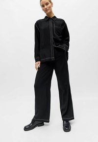 Femme portant une tenue noire ample avec des détails de couture blanche, debout avec une main dans la poche et des chaussures noires sur un fond blanc.