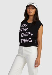 Black sleeveless sweater with "JOY OVER EVERY THING" in pink text, ribbed neckline and hem. Worn with white pants, casual fit.