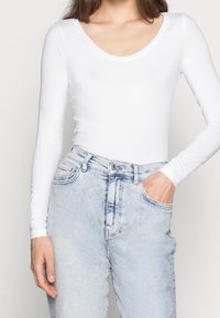 Kvinna i en figurnära vit långärmad topp som är instoppad i högmidjade ljusblå denimjeans, med ena handen i fickan, neutral bakgrund.