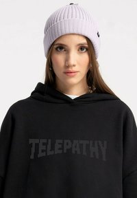 Fioletowy, żeberkowany beanie z zaokrąglonym czubkiem, zestawiony z czarną bluzą z kapturem, na której znajduje się tekst "TELEPATHY" w wyraźnym, pikselowym stylu.