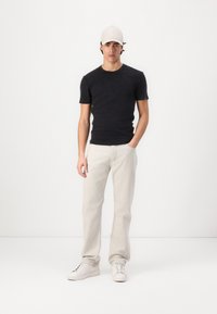 Sort kortærmet t-shirt, lys beige jeans og hvide sneakers; model iført en lysegrå cap, stående på en hvid baggrund.