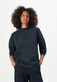 Pulover tricotat ribbat de culoare albastru închis, cu guler înalt și mâneci raglan. Are o croială lejeră și un material texturat. Asortat cu pantaloni de culoare albastră.