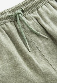 Gros plan sur un pantalon en tissu texturé vert clair avec une taille élastique et un cordon de serrage noué doté d'embouts métalliques argentés.