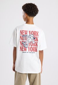 Valkoinen ylisuurikokoinen t-paita, jossa on sarjakuvahahmo ja punainen "NEW YORK" -teksti. Lyhyet hihat, pyöreä kaula-aukko, pehmeää puuvillakangasta.