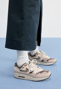 Nike Air Max 1 sportbačiai turi kreminės ir gyvačių rašo dizainą su tekstūruota viršutine dalimi, baltais raišteliais ir matomais oro pagalvėlėmis, derinami su baltais kojinėmis.