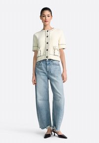 Mujer de pie que lleva un cárdigan corto de manga corta color crema con costuras y botones negros, vaqueros de pierna ancha azul claro y tacones puntiagudos negros.