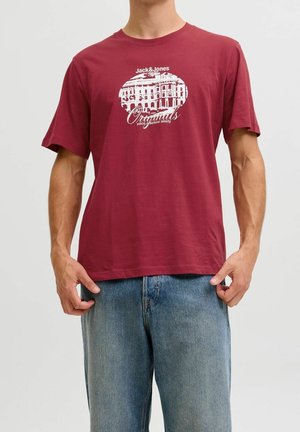Man wearing a maroon Jack & Jones Originals t-shirt with a white building graphic and text, paired with blue denim jeans.