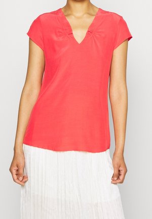 Kobieta ubrana w koralową bluzkę z krótkim rękawem i dekoltem w serek oraz białą plisowaną spódnicę, stojąca z ramionami swobodnie opuszczonymi wzdłuż ciała.