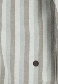 Pasiasta tkanina w jasnoszarym i beżowym kolorze o miękkiej teksturze; zawiera okrągły ciemny guzik z wytłoczonym wzorem.