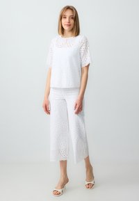 Top bianco ricamato a occhiello e pantaloni cropped abbinati, con bordi ondulati e ritagli circolari, caratterizzati da una consistenza liscia e una vestibilità rilassata.