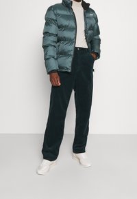 Uomo che indossa un giubbotto trapuntato teal, un dolcevita crema, pantaloni in velluto a coste scuri e sneakers bianche, in piedi contro un muro semplice.
