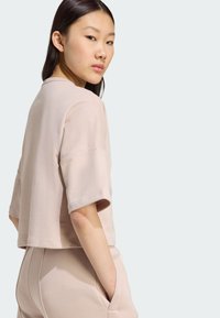 Svijetloružičasti skraćeni dukserica s kratkim rukavima, mekane teksture i opuštenog kroja, uparen s odgovarajućim hlačama. Vidljiv detalj džepa.
