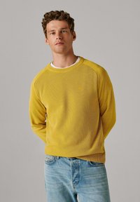 Kollane crew kaelusega pusa tekstuurse kangaga, raglaanvarrukatega ja peene logodetailiga, kombineeritud helesiniste teksadega.