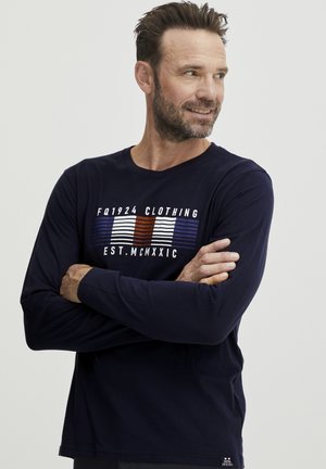 Marineblauwe longsleeve shirt van katoen met een ronde hals. Voorzien van witte en rode tekst en strepen aan de voorkant, en een klein logo op de zoom.