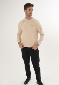 Helles Pfirsich-Sweatshirt mit Raglanärmeln, kleines braunes Logo auf der Brust, kombiniert mit schwarzen Jeans und schwarzen Sneakers, vor einem weißen Hintergrund.