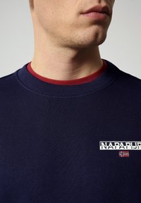 Granatowy sweter z ribbowym kołnierzem i bordowym wykończeniem wewnętrznym. Posiada białe logo "NAPAPIJRI" oraz akcent w postaci norweskiej flagi.
