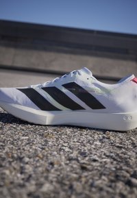 Weißer Laufschuh mit schwarzen Streifen und rotem Detail an der Ferse, ruhend auf einem Kiesuntergrund mit Betonhintergrund.