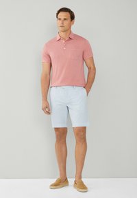 Man i en rosa kortärmad polo, ljusblå shorts och beige slipskor, stående mot en enkel ljusgrå bakgrund.