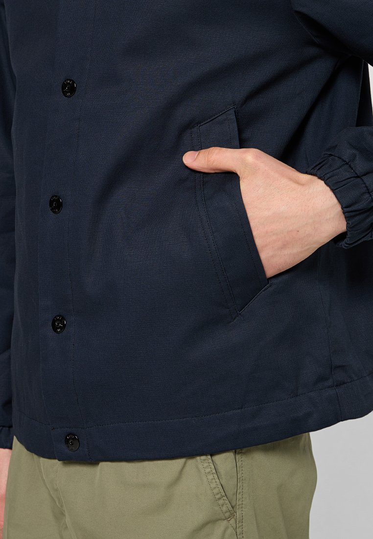 Navyblauwe jas met zwarte knopen, elastische manchetten en een zijzak. Gemaakt van textuurstof. Eenvoudig, klassiek ontwerp.