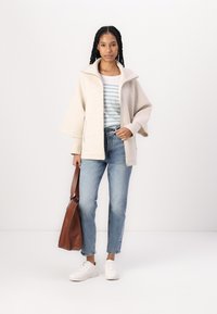 Lys beige oversized jakke med høj krave, suppleret med en stribet top, blå jeans og hvide sneakers. Bærer en brun skuldertaske.