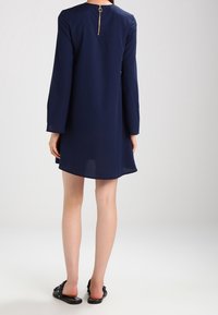 Marinblå klänning med långa ärmar, rund halsringning och en gulddetalj med dragkedja på baksidan. Slät tyg och längd ovanför knäna.