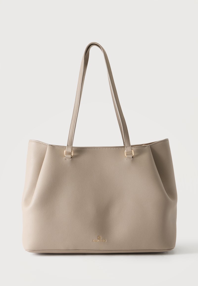 Sac cabas en cuir beige avec longues bretelles et logo doré discret à l’avant, sur un fond clair uni.