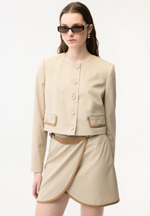 Donna che indossa una giacca beige con bottoni e tasche a patta, gonna a portafoglio coordinata con cintura, orecchini a cerchio dorati e occhiali da sole rettangolari scuri.