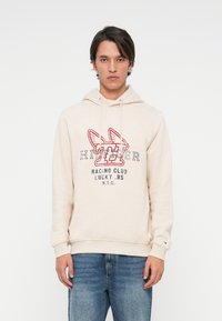 Młody mężczyzna w beżowej bluzie z kapturem z czerwono-czarnym grafiką "Hilfiger Racing Club Lucky .85 NYC", zestawionej z niebieskimi dżinsami, stojący na białym tle.