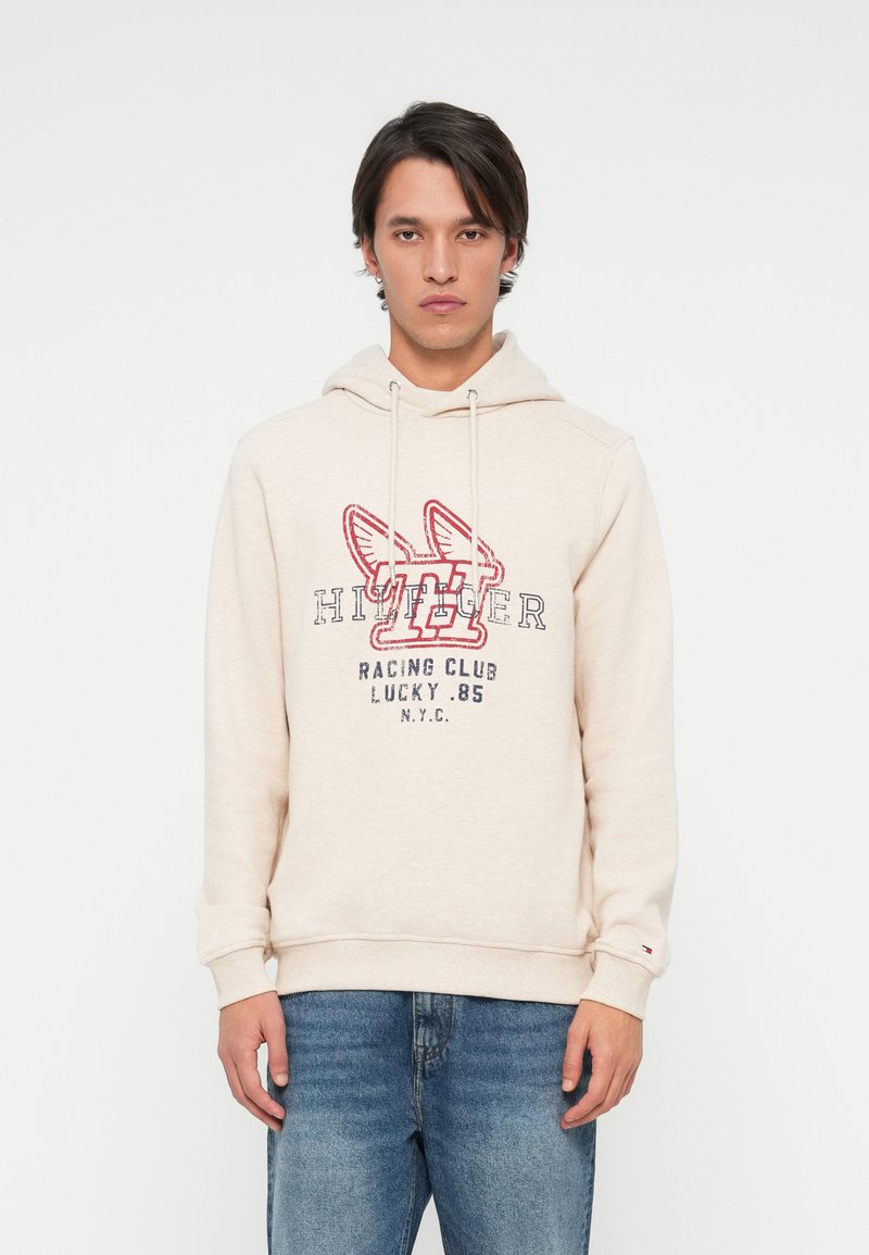 Młody mężczyzna w beżowej bluzie z kapturem z czerwono-czarnym grafiką "Hilfiger Racing Club Lucky .85 NYC", zestawionej z niebieskimi dżinsami, stojący na białym tle.