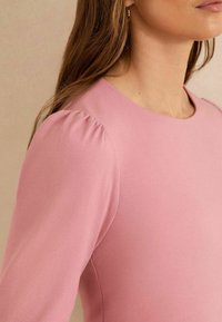 Rozā puff-sleeve tops izgatavots no gluda auduma, ar apaļu kakla izgriezumu un izsmalcinātām sānu šuvēm. Materiāls ir ar mīkstu tekstūru un nelielu spīdumu.