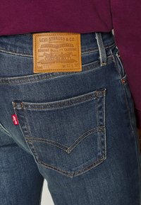 Jean en denim bleu foncé avec une étiquette en cuir arborant un logo embossé, des coutures dorées, et une petite étiquette rouge Levi's sur la poche arrière.
