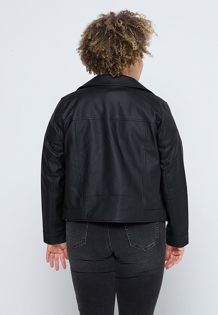 Persona con capelli ricci che indossa una giacca di pelle nera e jeans scuri, vista da dietro su uno sfondo semplice.