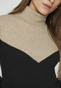 Pull en maille col roulé avec une partie supérieure beige et une partie inférieure noire, arborant un motif en chevrons, une texture douce et un col côtelé.