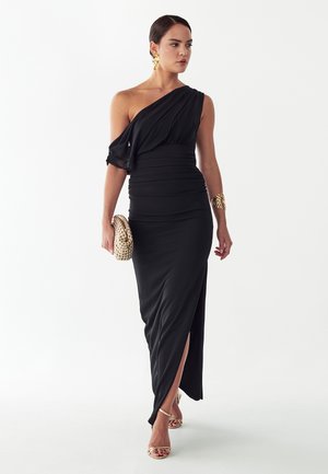 Vrouw in een zwarte galajurk met één schouder, geplooid en met een split langs de dij, houdt een beige gevlochten clutch vast, draagt gouden sieraden en sandalen met bandjes.