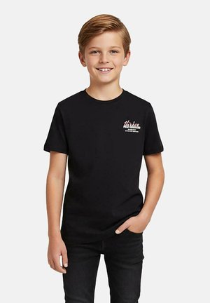 Glunderende blonde jongen draagt een zwart t-shirt met grafisch ontwerp en zwarte broek, staat met één hand in de zak tegen een effen achtergrond.