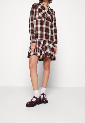 Kobieta w długiej sukience w kratę w kolorach czerwonym, czarnym i białym z falbaną u dołu, białe skarpetki do kostek oraz błyszczące bordowe, masywne buty.