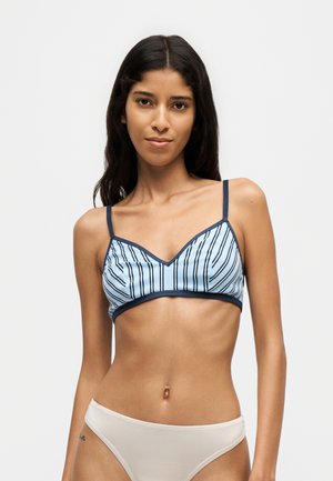 Donna con capelli lunghi e scuri indossa un reggiseno a righe blu e bianche con bordatura scura e biancheria intima beige chiaro su uno sfondo chiaro uniforme.