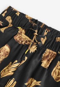 Pantaloni scurți negri cu textură lucioasă și talie elastică. Prezintă imprimeuri grafice aurii cu mâini și chipuri. Include un șnur cu accente galbene.