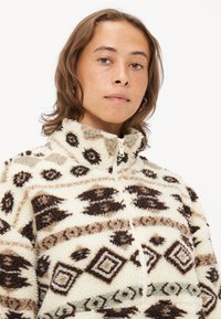 Fleece zip-up jakna v krem barvi z rjavimi tribalnimi vzorci. Ima visok ovratnik, teksturirano blago in kontrastno zadrgo.