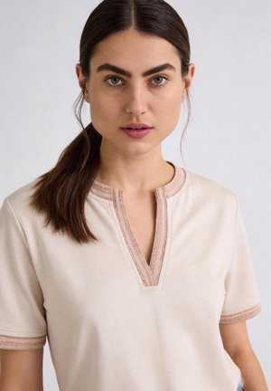 Junge Frau mit dunklen Haaren in einem tiefen Pferdeschwanz, die ein beiges Kurzarmoberteil mit rosa Besatz am Ausschnitt und an den Ärmeln trägt.