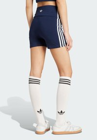 Námorné šortky s bielymi bočnými pruhmi, vysoký pás, kombinované s bielymi ponožkami po kolená s čiernymi pruhmi a logom Adidas, nosené s bielymi teniskami.