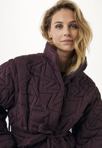 Donna bionda con occhi azzurri che indossa una giacca trapuntata viola legata in vita, che guarda direttamente nella fotocamera con un'espressione neutra.