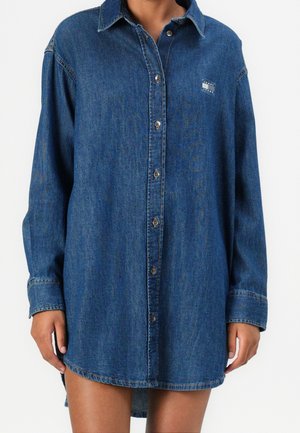 Langærmet blå denimskjortekjole med knapper foran og lille Tommy Jeans-logo på brystet, båret af en person, der står med afslappede arme.