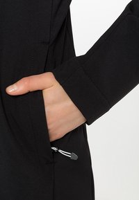 Main partiellement insérée dans la poche d'une veste noire avec des coutures visibles et une petite tirette de fermeture éclair noir et blanc.