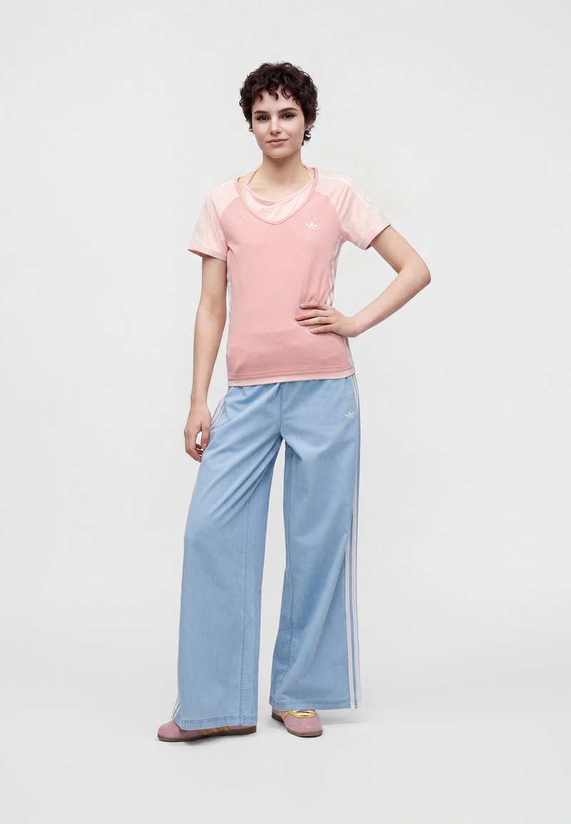Jeune personne debout, main sur la hanche, portant un haut rose à manches courtes et un pantalon ample bleu clair avec des bandes latérales et des baskets.